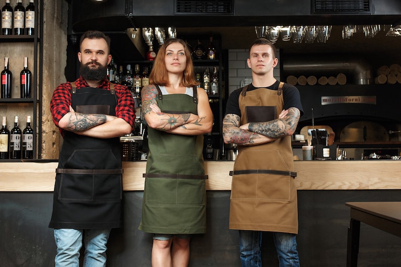 May include: Three people wearing different colored aprons stand in front of a bar counter. The person on the left is wearing a black apron with a brown leather strap, the person in the middle is wearing a green apron with a brown leather strap, and the person on the right is wearing a brown apron with a brown leather strap. All three people have tattoos on their arms.