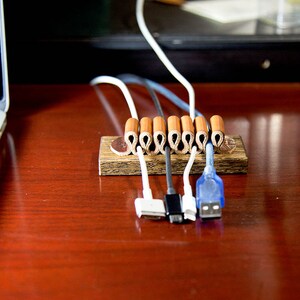 Puede incluir: Un organizador de cables de madera con seis envoltorios de cable de cuero marr&oacute;n. El organizador tiene cinco cables que pasan a trav&eacute;s de &eacute;l, incluyendo un cable blanco, un cable gris, un cable azul y dos cables blancos. Los cables est&aacute;n conectados a varios dispositivos electr&oacute;nicos.