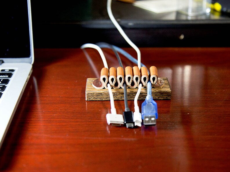 Puede incluir: Un organizador de cables de madera con seis envoltorios de cable de cuero marr&oacute;n. El organizador tiene cinco cables que pasan a trav&eacute;s de &eacute;l, incluyendo un cable blanco, un cable gris, un cable azul y dos cables blancos. Los cables est&aacute;n conectados a varios dispositivos electr&oacute;nicos.