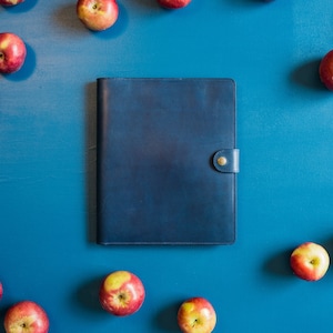 May include: A dark blue leather bound journal or notebook with a brass button closure. The journal is surrounded by red apples on a blue background.