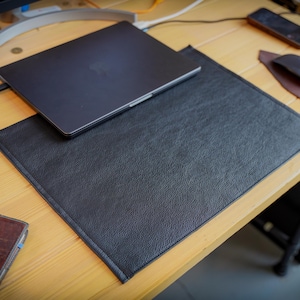 Könnte beinhalten: Eine rechteckige schwarze Schreibtischunterlage mit strukturierter Oberfläche und genähten Kanten. Die Unterlage liegt auf einer Holzoberfläche, teilweise unter einem geschlossenen dunklen Laptop, und bietet eine flache Schutzfläche.