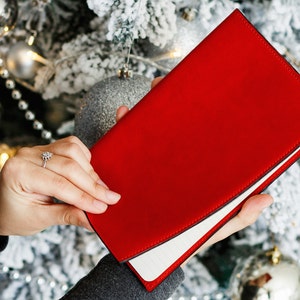 Puede incluir: Un diario de cuero rojo con interior blanco, sostenido en una mano sobre un fondo de un árbol de Navidad decorado con adornos plateados.