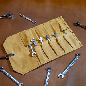 May include: A tan canvas roll-up tool holder with a leather strap. The holder has eight slots for wrenches. The holder is open and several wrenches are visible. There are also several wrenches scattered around the holder on a brown surface.