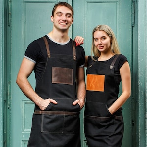 May include: Two people wearing black aprons with brown leather accents and pockets. The aprons have adjustable straps. The people are standing in front of a green door.
