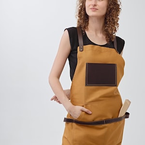 May include: A woman wearing a brown canvas apron with a brown leather pocket and a brown leather strap. She is standing with her hands in her pockets and looking up.