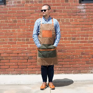 May include: A person wearing a blue and white plaid shirt and a brown leather and canvas apron with a green pocket. The apron has a brown leather strap and buckle. The person is standing in front of a brick wall.