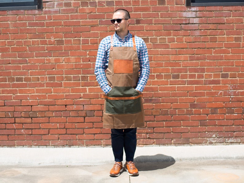 May include: A person wearing a blue and white plaid shirt and a brown leather and canvas apron with a green pocket. The apron has a brown leather strap and buckle. The person is standing in front of a brick wall.