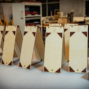 Puede incluir: Una colección de expositores de madera con acabado claro. Cada expositor tiene forma rectangular con detalles oscuros en las esquinas y un emblema decorativo en la parte superior. Los expositores están dispuestos en fila, mostrando un diseño minimalista.