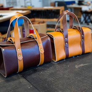 K&ouml;nnte beinhalten: Zwei Ledertaschen mit braunen und braunen Lederriemen und Messingnieten. Die Tasche links ist dunkelbraun und die Tasche rechts ist braun.