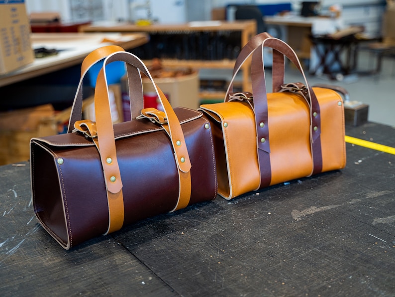 K&ouml;nnte beinhalten: Zwei Ledertaschen mit braunen und braunen Lederriemen und Messingnieten. Die Tasche links ist dunkelbraun und die Tasche rechts ist braun.