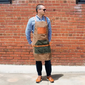 May include: A person wearing a brown leather and canvas apron with multiple pockets and a green canvas pocket. The apron has brown leather straps and a brown leather patch on the front. The person is standing in front of a brick wall.