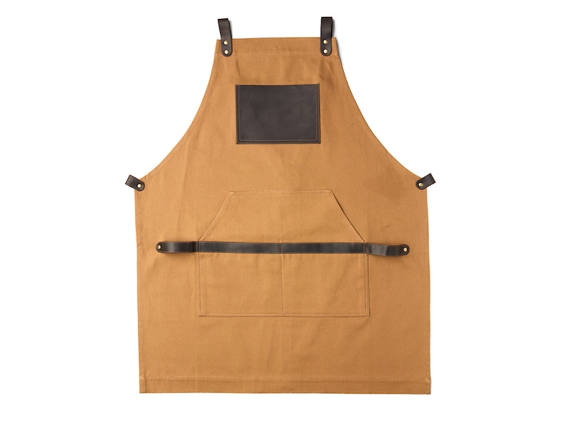 May include: A brown canvas apron with a large brown leather patch on the front. The apron has a large pocket on the front and adjustable straps.