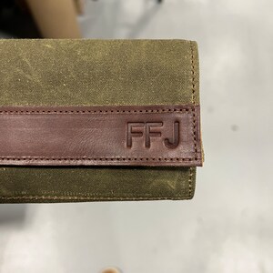 May include: A green canvas wallet with a brown leather flap. The flap has the initials "FFJ" embossed in it.