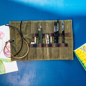 K&ouml;nnte beinhalten: Eine gr&uuml;ne Canvas-Rolltasche mit Lederriemen und einem braunen Ledergriff. Die Tasche ist gef&uuml;llt mit verschiedenen Kunstbedarfsartikeln, darunter Pinsel, Bleistifte und Stifte.