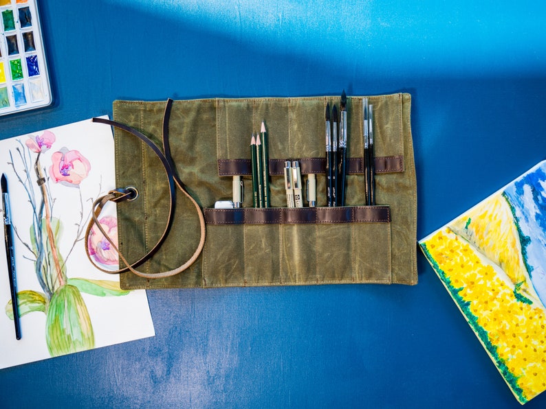 K&ouml;nnte beinhalten: Eine gr&uuml;ne Canvas-Rolltasche mit Lederriemen und einem braunen Ledergriff. Die Tasche ist gef&uuml;llt mit verschiedenen Kunstbedarfsartikeln, darunter Pinsel, Bleistifte und Stifte.