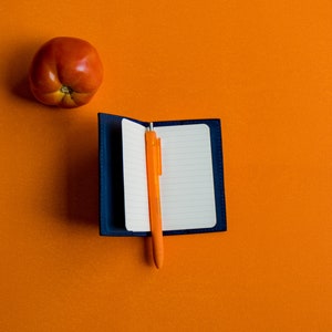 May include: A blue notebook with a blank white page and an orange pen on an orange background. A red tomato is in the upper left corner of the image.