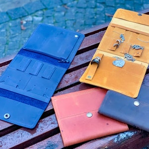 May include: A collection of leather jewelry organizers in various colors, including blue, orange, red, and brown. The organizers are open and closed, showcasing compartments for earrings, necklaces, and rings. Some organizers hold jewelry.