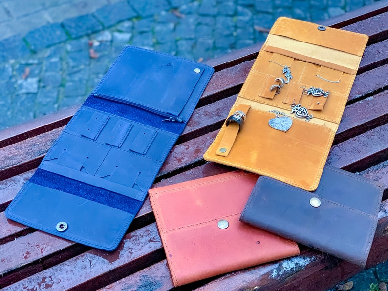 May include: A collection of leather jewelry organizers in various colors, including blue, orange, red, and brown. The organizers are open and closed, showcasing compartments for earrings, necklaces, and rings. Some organizers hold jewelry.