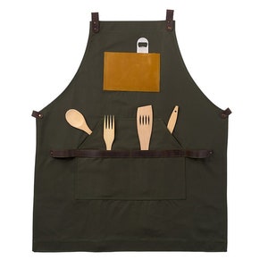 May include: A dark green canvas apron with brown leather straps and a brown leather pocket. The pocket has a small, light brown leather patch on the front. The apron has a bottle opener attached to the top strap. There are four wooden utensils in the pocket: a spoon, a fork, a spatula, and a knife.