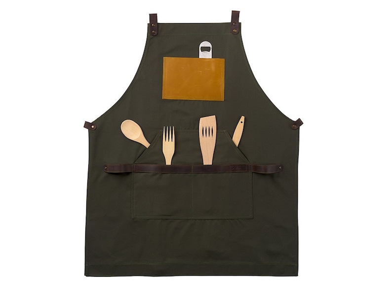 May include: A dark green canvas apron with brown leather straps and a brown leather pocket. The pocket has a small, light brown leather patch on the front. The apron has a bottle opener attached to the top strap. There are four wooden utensils in the pocket: a spoon, a fork, a spatula, and a knife.