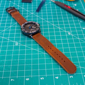 Op de afbeelding: Een bruin leren horlogebandje met een zwarte wijzerplaat en blauwe accenten. Het horloge ligt op een groene snijmat met witte markeringen.