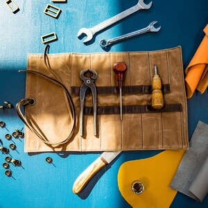 May include: A brown leather tool roll with a variety of tools, including pliers, awls, and a knife. The roll is unrolled and laid flat on a blue surface. There are also various leather scraps, metal rivets, and other tools scattered around the roll.