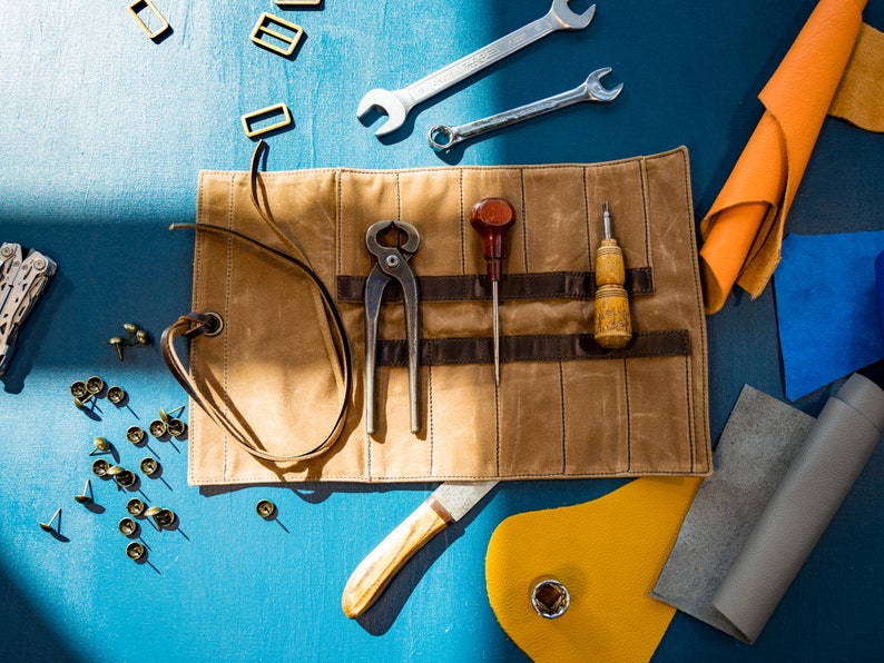 May include: A brown leather tool roll with a variety of tools, including pliers, awls, and a knife. The roll is unrolled and laid flat on a blue surface. There are also various leather scraps, metal rivets, and other tools scattered around the roll.