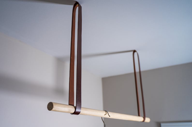 K&ouml;nnte beinhalten: Eine Holzstange h&auml;ngt an zwei braunen Lederriemen, die von der Decke abgeh&auml;ngt sind. Die Stange ist etwa 76 cm lang.