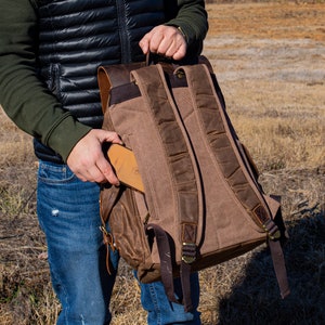 Puede incluir: Una mochila de lona marr&oacute;n con detalles de cuero. La mochila tiene correas ajustables, m&uacute;ltiples bolsillos y un asa superior. Se est&aacute; colocando una cartera de cuero marr&oacute;n en el bolsillo lateral. La mochila est&aacute; dise&ntilde;ada para uso en exteriores.