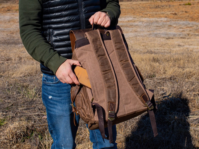 Puede incluir: Una mochila de lona marr&oacute;n con detalles de cuero. La mochila tiene correas ajustables, m&uacute;ltiples bolsillos y un asa superior. Se est&aacute; colocando una cartera de cuero marr&oacute;n en el bolsillo lateral. La mochila est&aacute; dise&ntilde;ada para uso en exteriores.
