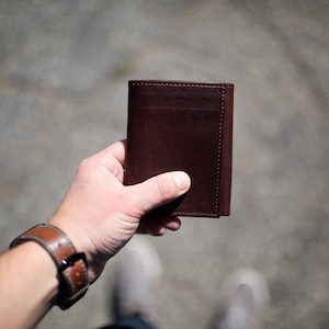May include: A dark brown leather wallet with a minimalist design. The wallet is held in a hand, showcasing its compact size and smooth texture. The leather appears to be high-quality, with visible stitching along the edges.
