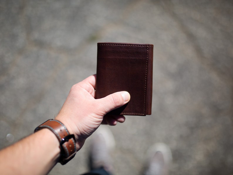 May include: A dark brown leather wallet with a minimalist design. The wallet is held in a hand, showcasing its compact size and smooth texture. The leather appears to be high-quality, with visible stitching along the edges.