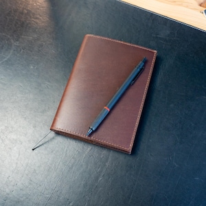 May include: A brown leather journal with a black pen resting on top. The pen has a red band near the tip. The journal is on a dark surface, possibly a desk or table. The scene suggests a workspace or study area.