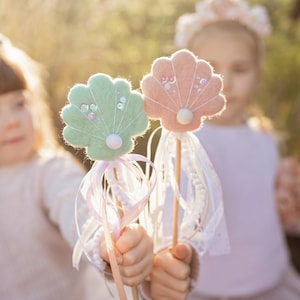 Peut inclure: Deux enfants tiennent des baguettes en forme de coquillages en feutre de couleur pastel avec un ruban blanc et des accents de perles. Les baguettes sont sur des bâtonnets en bois.