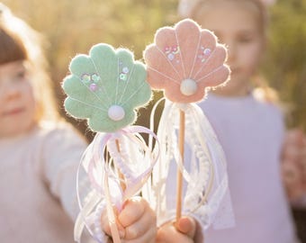 Puede incluir: Dos niños sostienen varitas de conchas marinas de fieltro de color pastel con cinta blanca y detalles de perlas. Las varitas están en palos de madera.