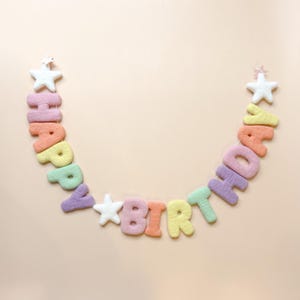 Può includere: Una ghirlanda in feltro che compone le parole HAPPY BIRTHDAY in colori pastello tra cui rosa, arancione, giallo, verde e viola. Lo striscione presenta tre stelle in feltro bianco, una a ciascuna estremità e una tra le due parole.