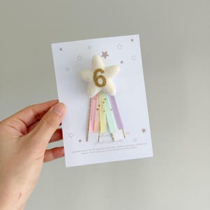 May include: A white card with a star-shaped embellishment featuring the number 6 in gold glitter. Below the star are pastel-colored ribbons in rainbow hues. The card has small star illustrations and text at the bottom.