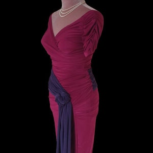Puede incluir: Un vestido largo de color fucsia ajustado con una faja morada anudada en un lado. El vestido tiene un escote en V y mangas cortas.