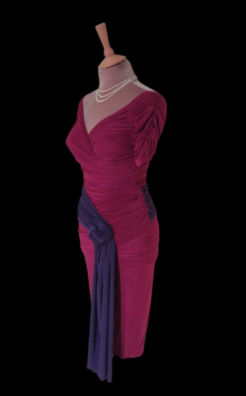 Puede incluir: Un vestido largo de color fucsia ajustado con una faja morada anudada en un lado. El vestido tiene un escote en V y mangas cortas.
