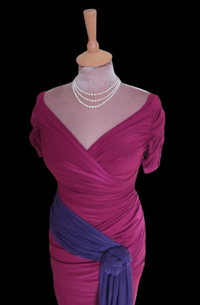 Puede incluir: Un vestido magenta con una faja morada anudada a la cintura. El vestido tiene un escote en V y mangas cortas. El vestido est&aacute; en un maniqu&iacute; y tiene un collar de perlas.