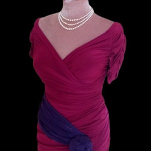 Puede incluir: Un vestido rosa oscuro sin mangas con una faja morada atada a la cintura. El vestido tiene un escote en V y una silueta ajustada.