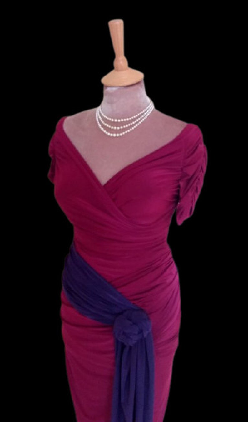 Puede incluir: Un vestido rosa oscuro sin mangas con una faja morada atada a la cintura. El vestido tiene un escote en V y una silueta ajustada.