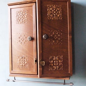 May include: A dark brown wooden cabinet with two doors and ornate carvings. The cabinet has a metal towel bar attached to the bottom.
