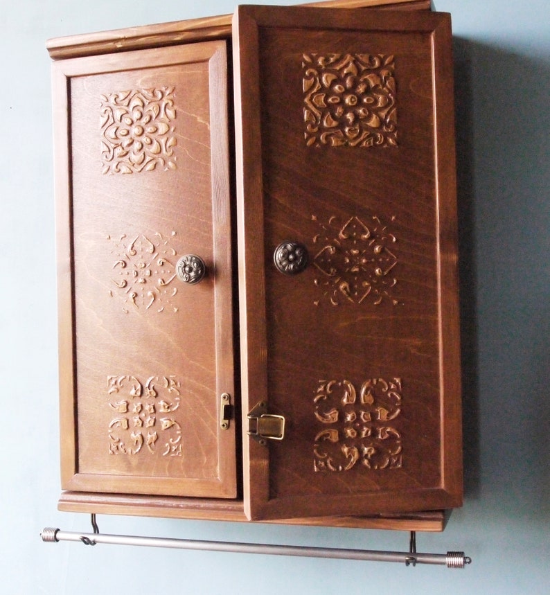 May include: A dark brown wooden cabinet with two doors and ornate carvings. The cabinet has a metal towel bar attached to the bottom.