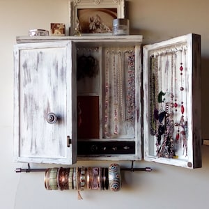 May include: White wooden jewelry cabinet with a distressed finish. The cabinet has a hinged door and a mirrored interior. The cabinet is filled with necklaces, earrings, and bracelets. There is a metal bar underneath the cabinet for hanging bracelets.