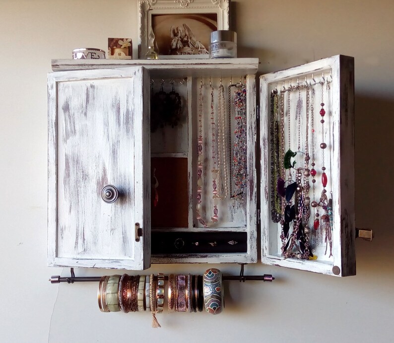 May include: White wooden jewelry cabinet with a distressed finish. The cabinet has a hinged door and a mirrored interior. The cabinet is filled with necklaces, earrings, and bracelets. There is a metal bar underneath the cabinet for hanging bracelets.
