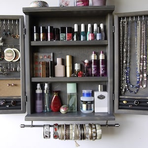 May include: A gray wooden jewelry cabinet with two doors that open to reveal shelves for storing jewelry, makeup, and other accessories. The cabinet has a pull-out rod for hanging bracelets.