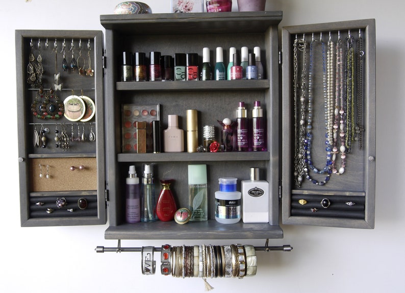May include: A gray wooden jewelry cabinet with two doors that open to reveal shelves for storing jewelry, makeup, and other accessories. The cabinet has a pull-out rod for hanging bracelets.