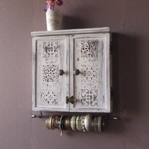 May include: White wooden cabinet with two doors featuring carved floral designs. The cabinet has a shelf and a metal rod underneath for hanging jewelry.