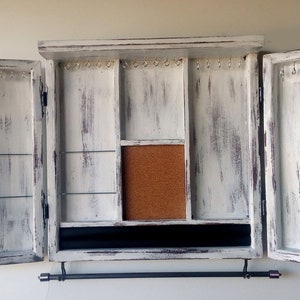 May include: White wooden jewelry cabinet with two doors open, revealing compartments for storing necklaces, earrings, and other jewelry. The cabinet has a distressed finish and a black metal rod for hanging necklaces.
